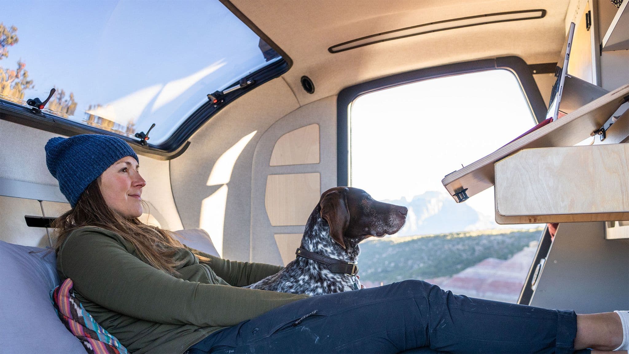 Woman in a beanie and green jacket lounges inside an Escapod trailer with her dog, enjoying a scenic mountain view through the open side door.