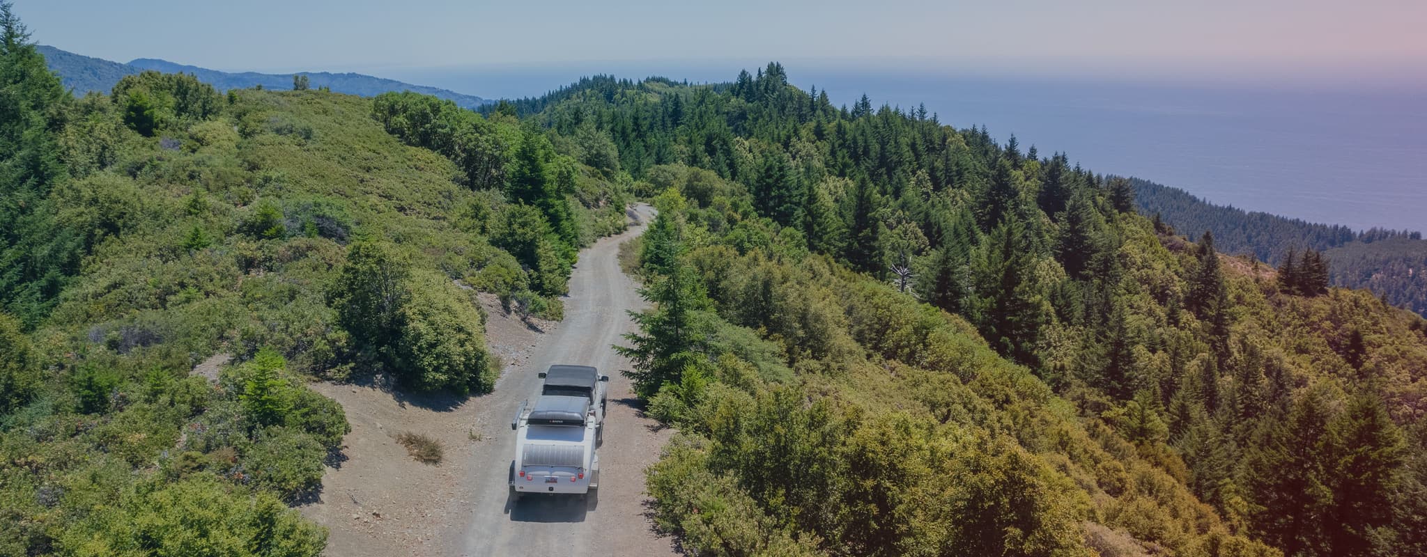 A Ford Bronco towing an Escapod trailer makes its way down a rugged dirt trail through mountainous terrain, surrounded by thick brush and pine trees, with the Pacific Ocean in the distance.