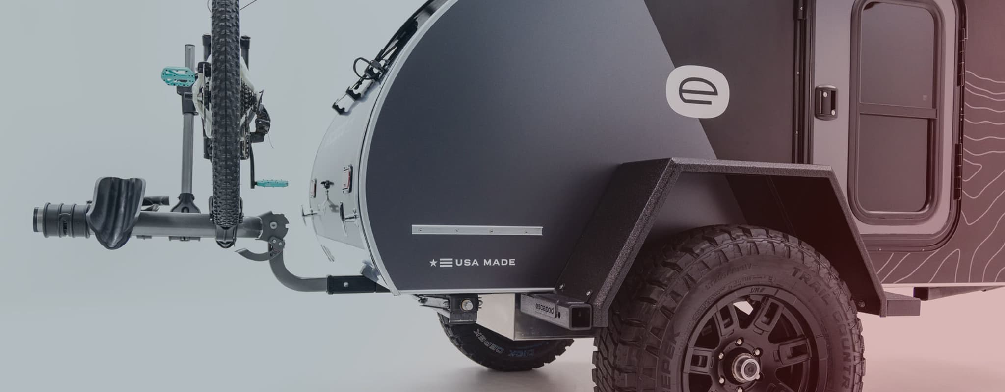 An Escapod Original TOPO, photographed in a studio environment, USA MADE sticker in the lower corner of the trailer, and a mountain bike on a rear hitch mounted rack.