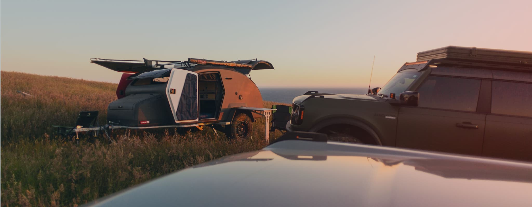 Escapod TOPO2 trailer parked on a grassy coastal hilltop at sunset, with vehicles and camping gear set up for an outdoor adventure.