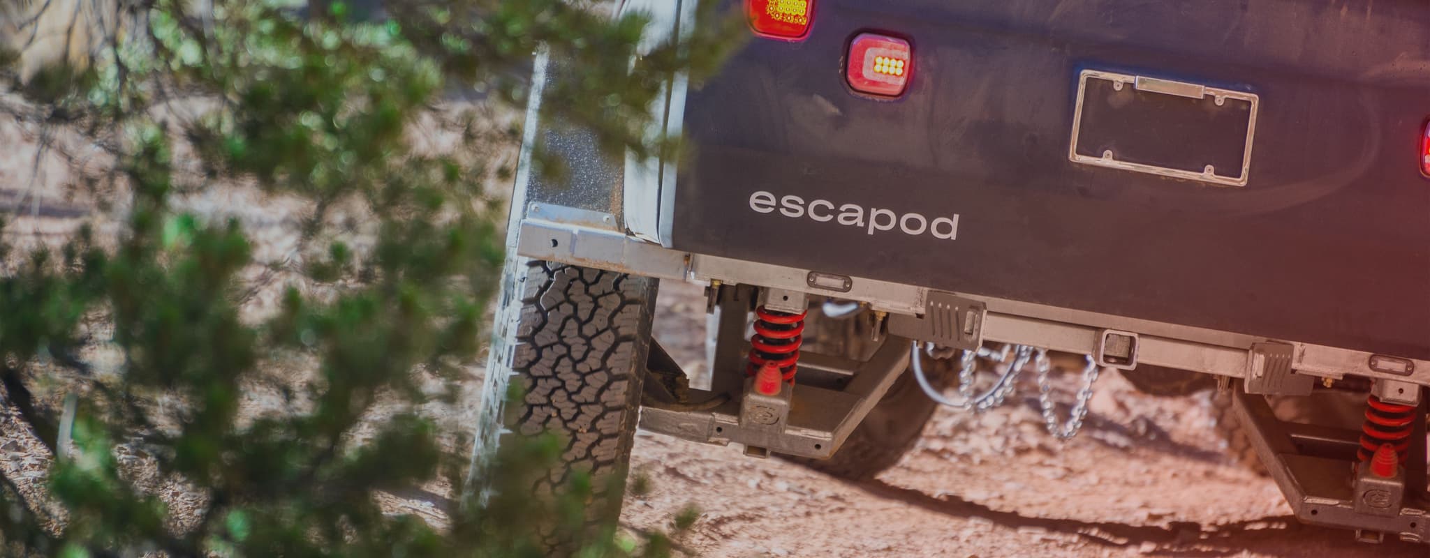 Close-up of an Escapod trailer’s suspension system and off-road tires, partially framed by desert foliage.