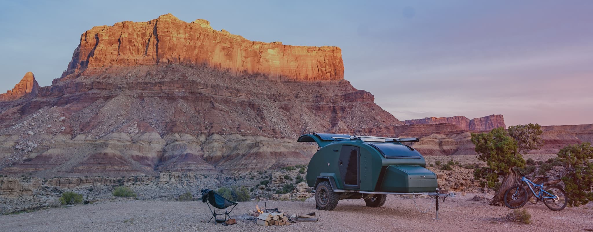 Escapod TOPO2 trailer parked at a scenic desert campsite near towering red rock formations, with a campfire, chair, and mountain bike nearby.