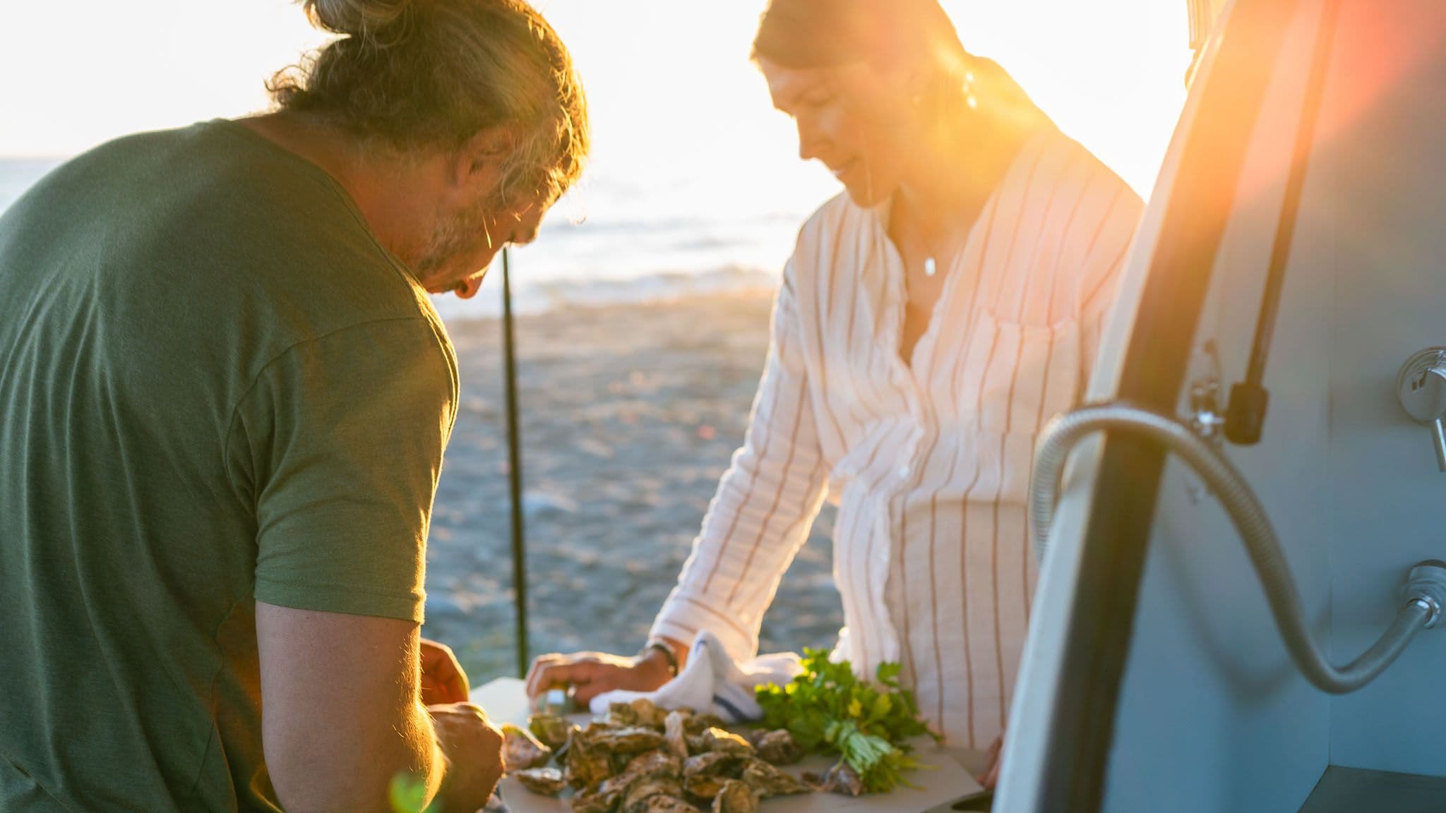 A man and woman prepare fresh oysters on a table next to an Escapod TOPO2 trailer, with the sun setting over the beach in the background.