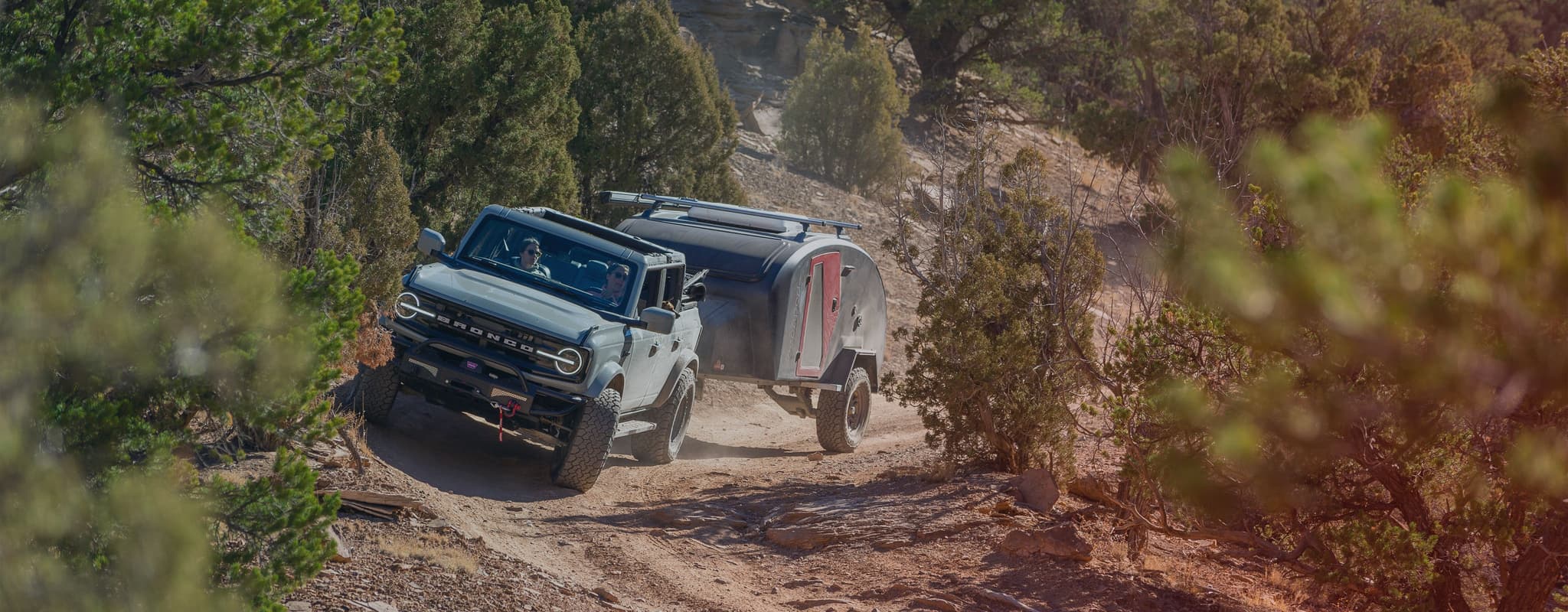 A Ford Bronco navigating a rugged dirt trail while towing an Escapod TOPO2 trailer, surrounded by dense desert vegetation.