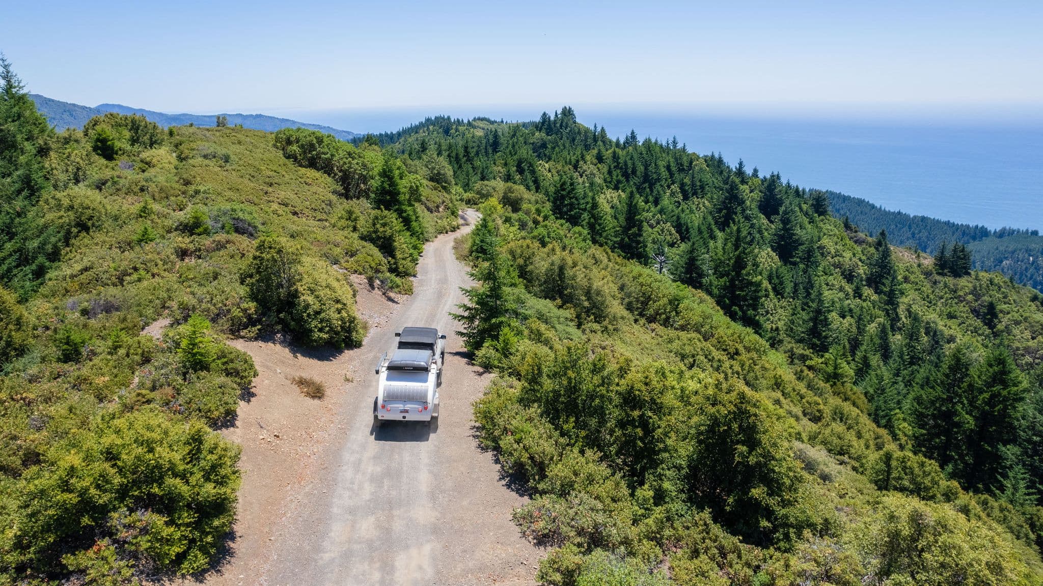 A white Escapod trailer and off-road vehicle drive along a scenic coastal dirt road, surrounded by rolling green hills and overlooking the Pacific Ocean.