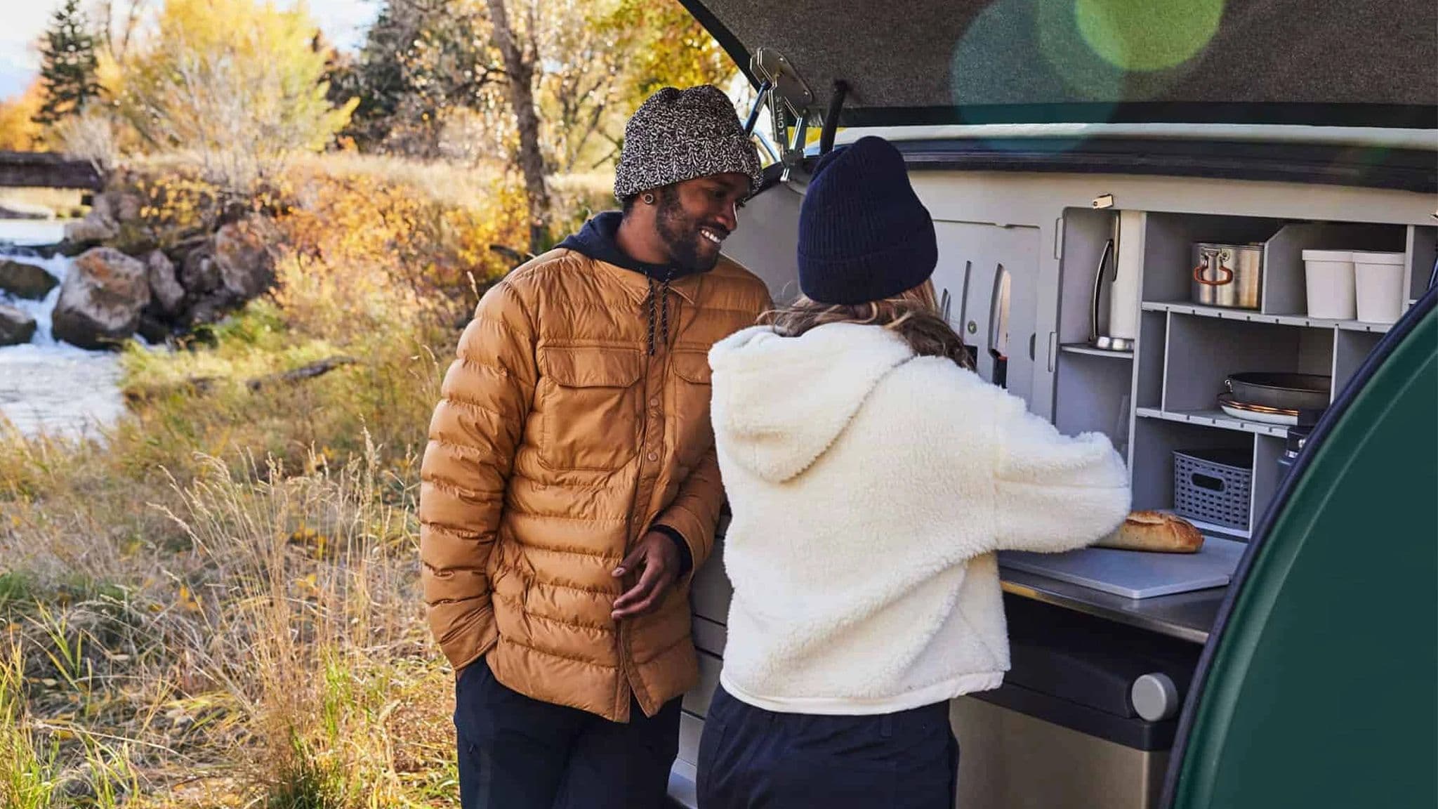 A couple preparing food at the open rear galley of an Escapod TOPO2 trailer, parked near a rushing creek with autumn foliage in the background.