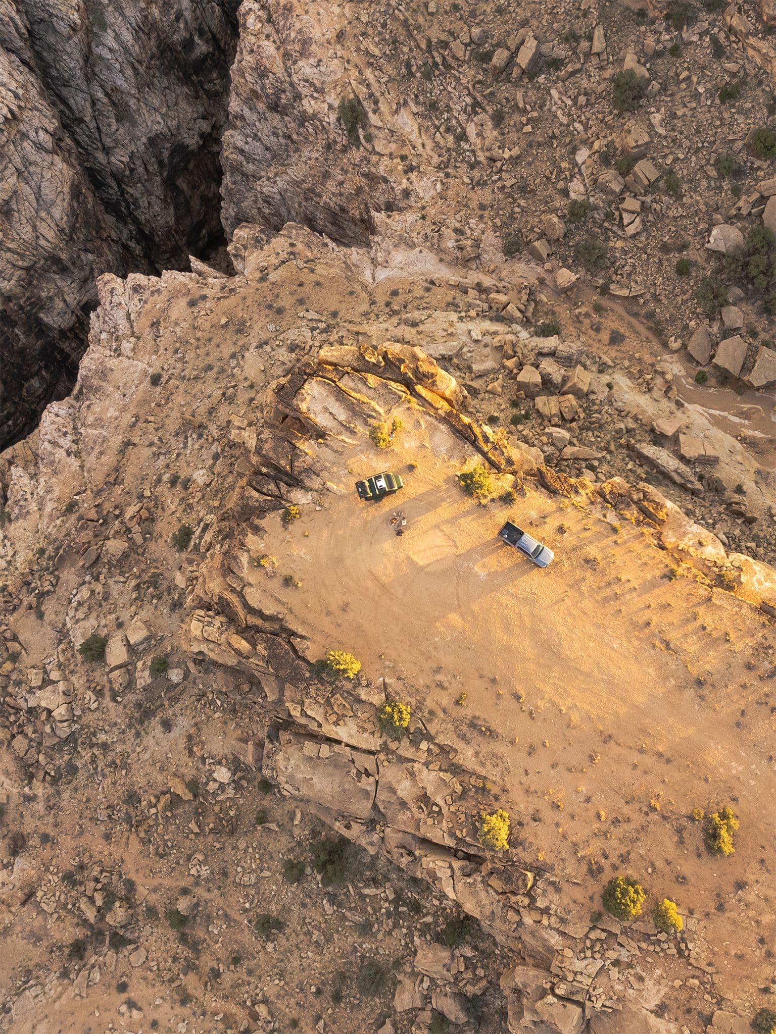 Aerial view of two off-road vehicles parked at the edge of a desert cliff, overlooking a deep canyon with golden light casting shadows.