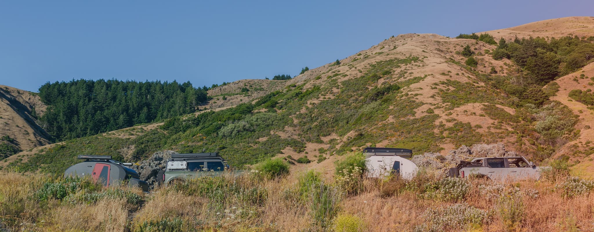 Two off-road camping trailers and vehicles set up in a remote, hilly landscape with dry grass, scattered bushes, and a forested ridge.