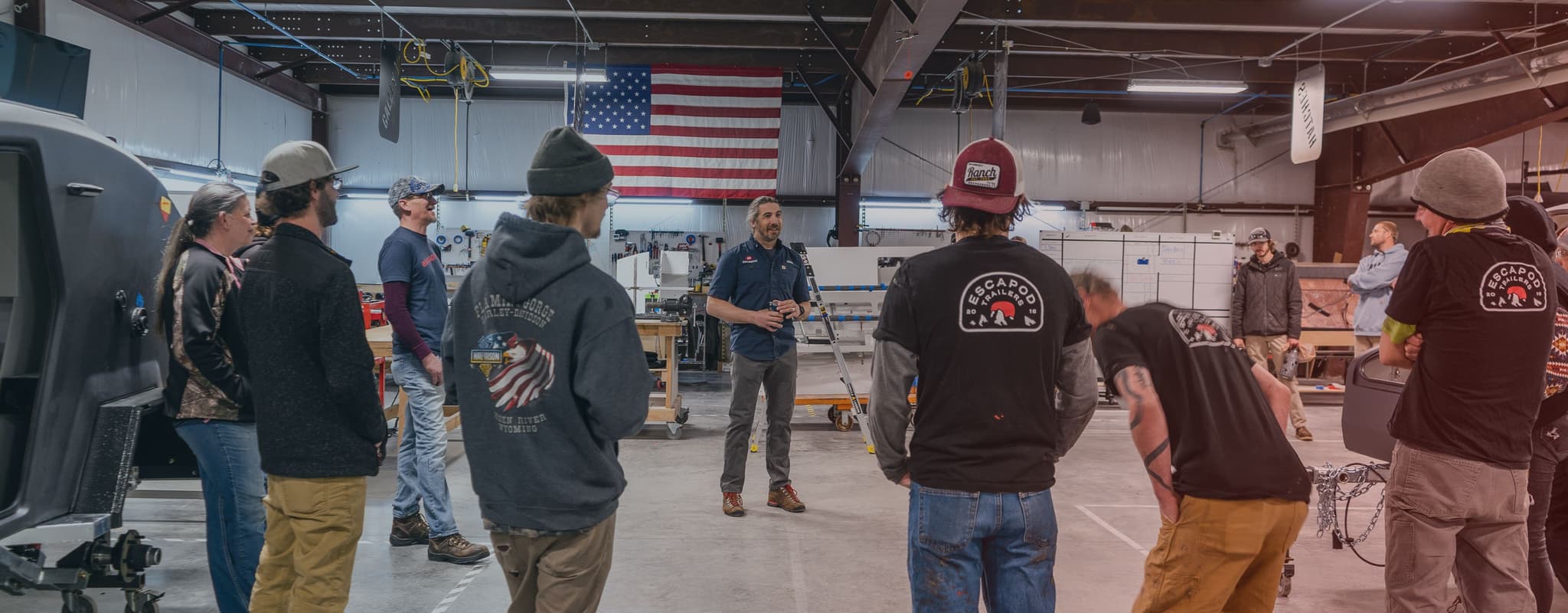 Escapod team meeting inside the production facility, employees gathered around a speaker, with an American flag displayed in the background.