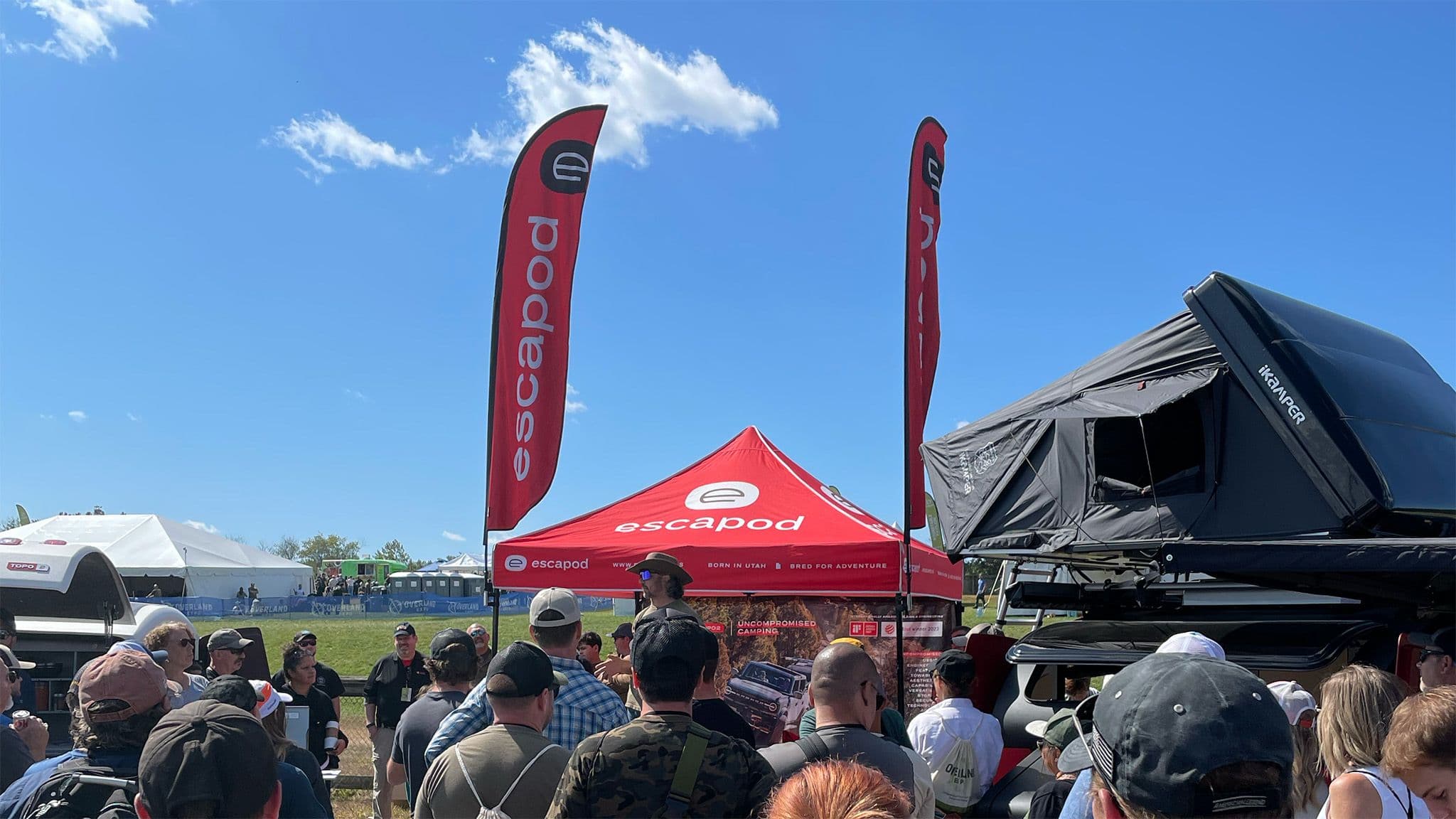 A red Escapod tent with two red Escapod flags at an overland event, with lots of event attendees in the foreground, an open iKamper tent on the right, and a blue sky with a solitary cloud.