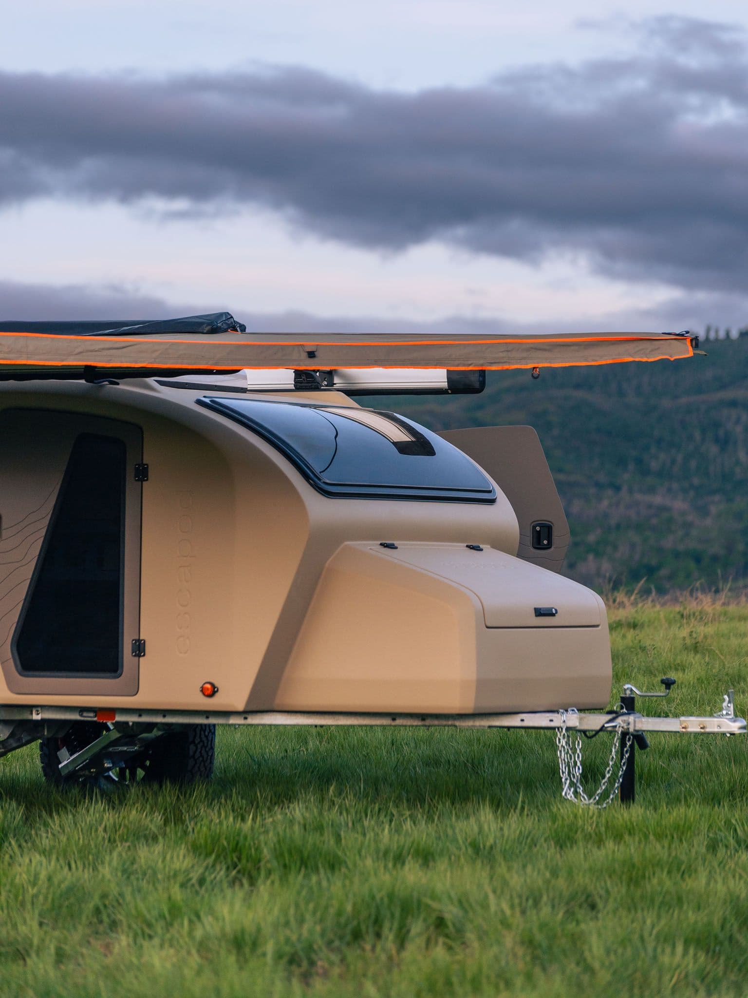 Side view of an Escapod TOPO2 trailer with an extended awning, set against a backdrop of dark clouds and distant mountains.
