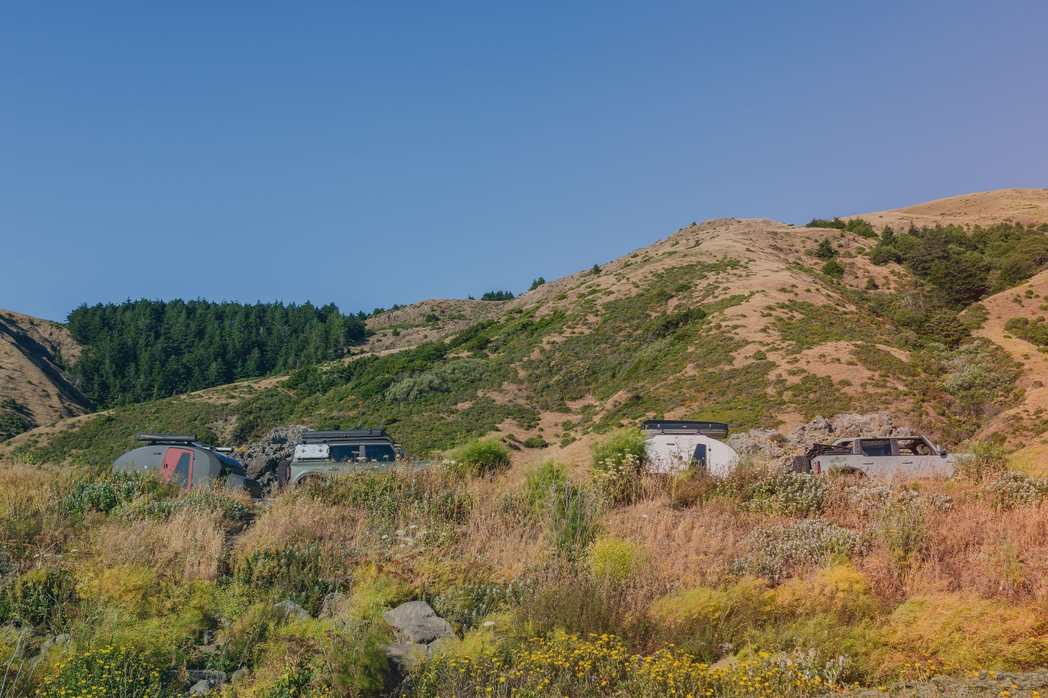 Two off-road camping trailers and vehicles set up in a remote, hilly landscape with dry grass, scattered bushes, and a forested ridge.