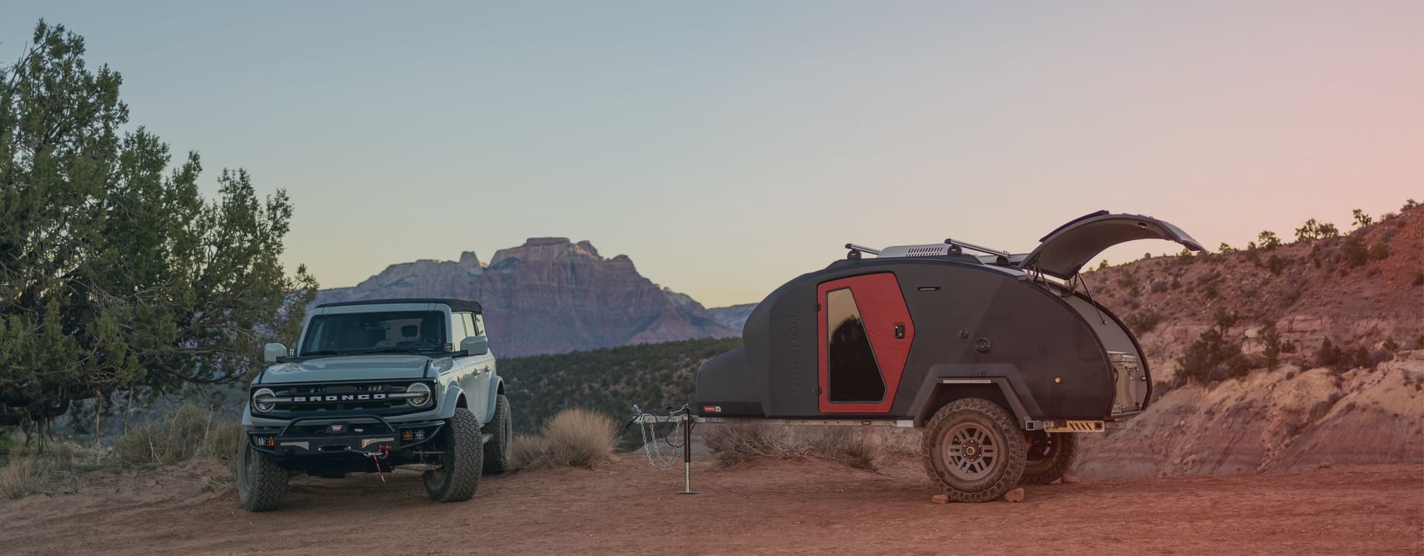 An Escapod TOPO2 trailer parked at a scenic overlook in the desert at sunset, hitched to a Ford Bronco with a mountain range in the background.
