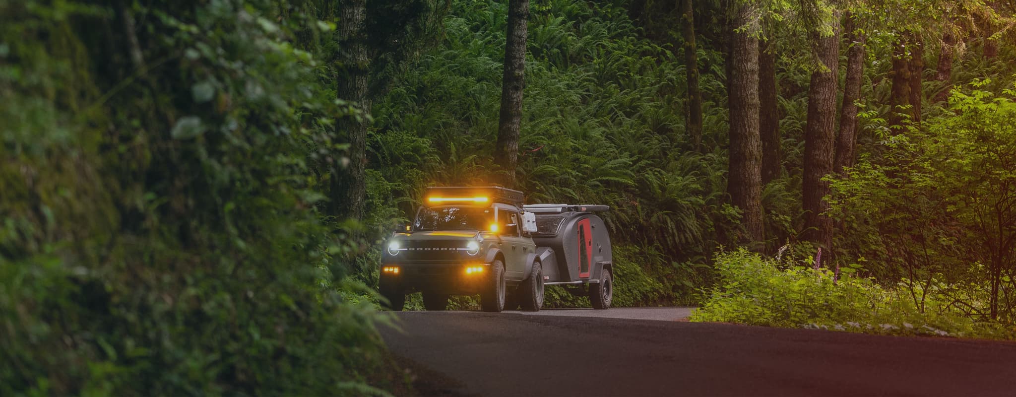 A rugged off-road vehicle towing an Escapod TOPO2 trailer through a dense, green forest, headlights and auxiliary lights glowing in the dim light.