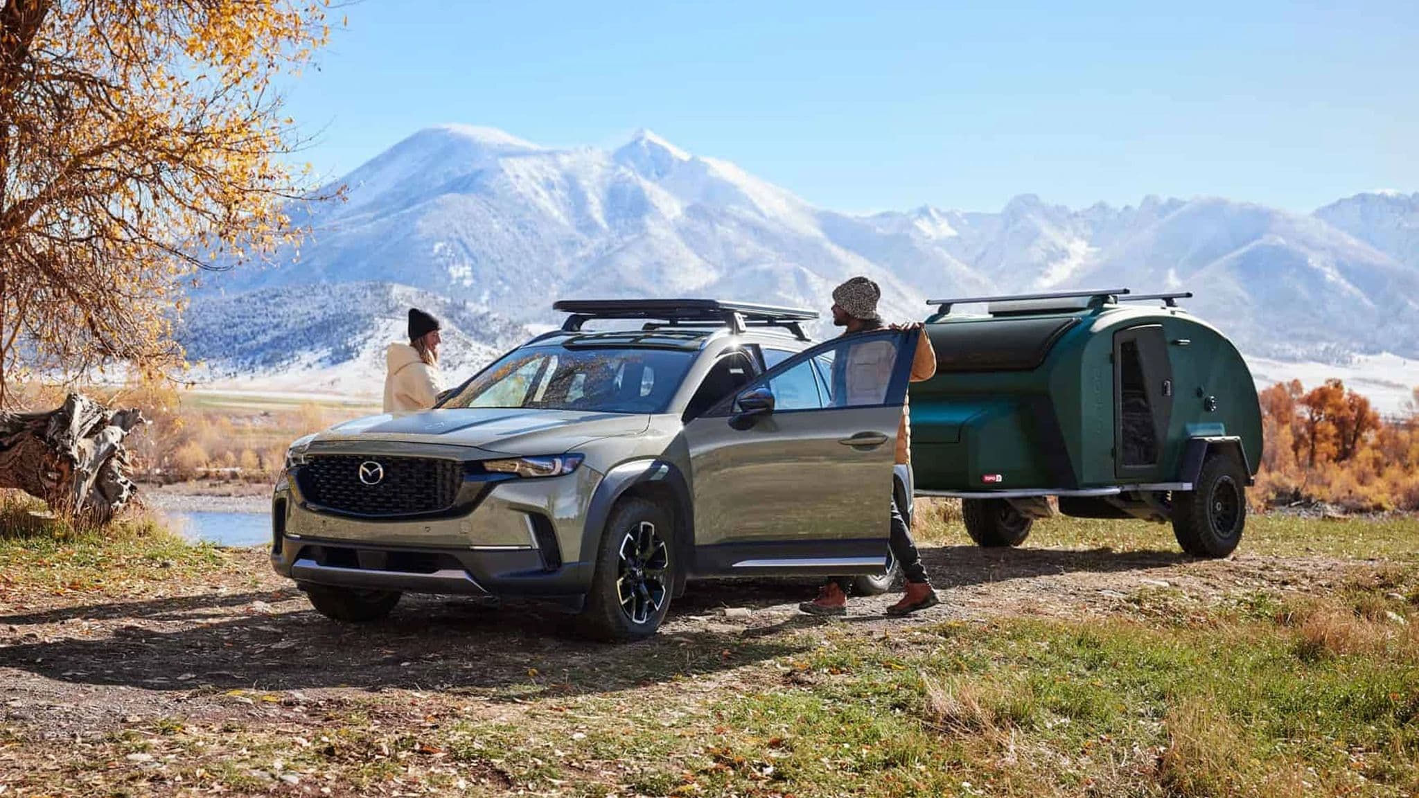 A couple arriving at a remote lakeside camping spot, stepping out of a Mazda CX-50 with an Escapod TOPO2 trailer parked nearby against a backdrop of snowy mountains and fall trees.