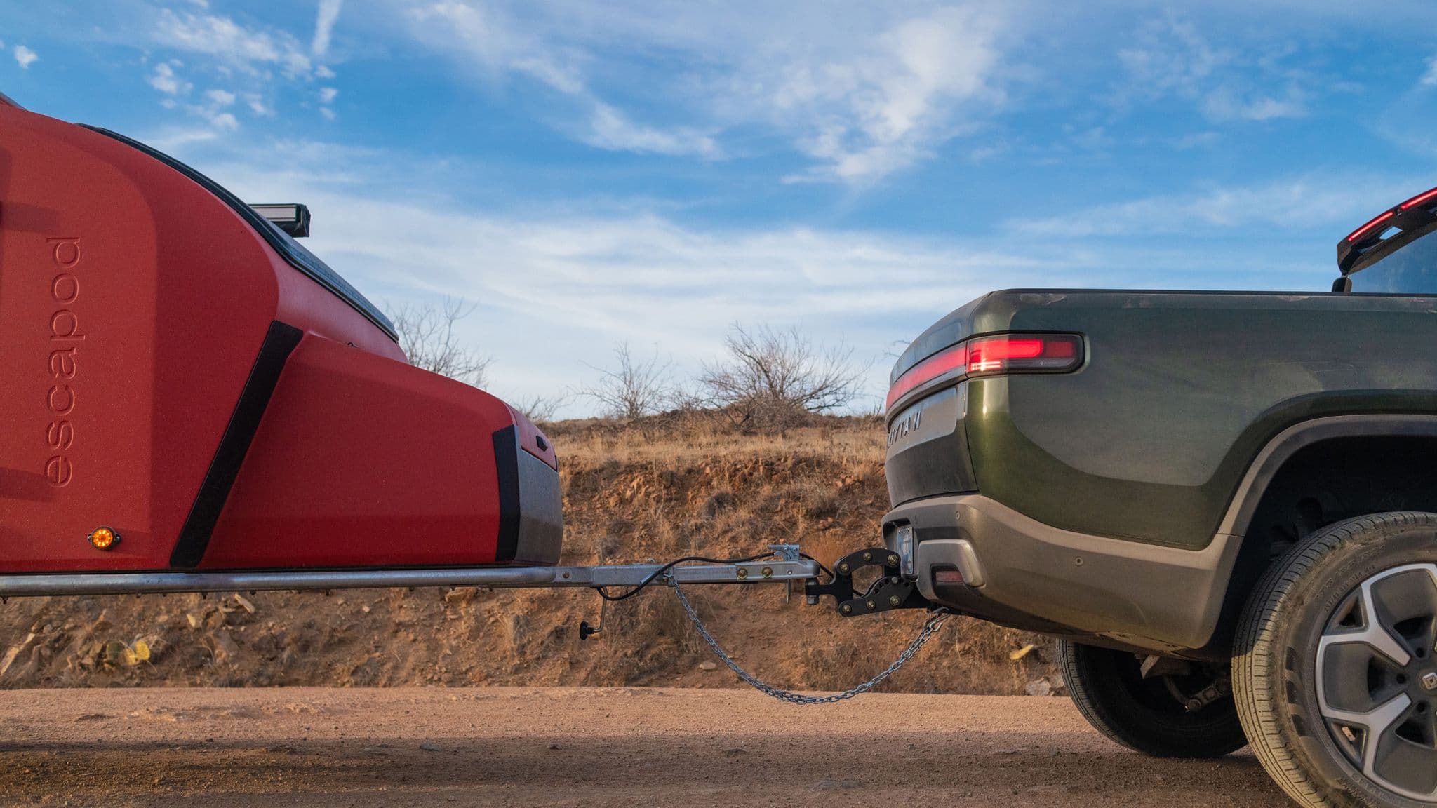 A hitch connection between a orange Escapod TOPO2 MTB trailer and a green Rivian truck on a dirt road, with rugged desert scenery in the background.