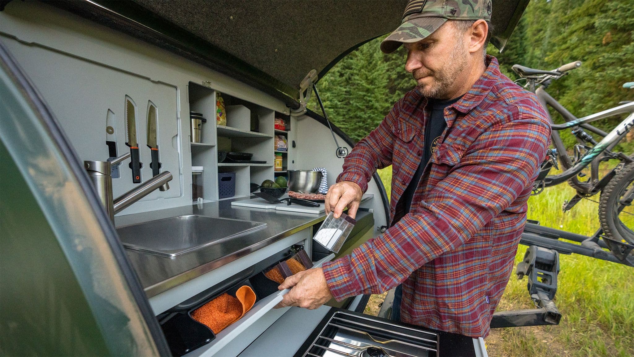 A man in a plaid shirt and a camouflage hat puts a spice container back into the spice rack, located below the sink in the galley of an Escapod TOPO2.