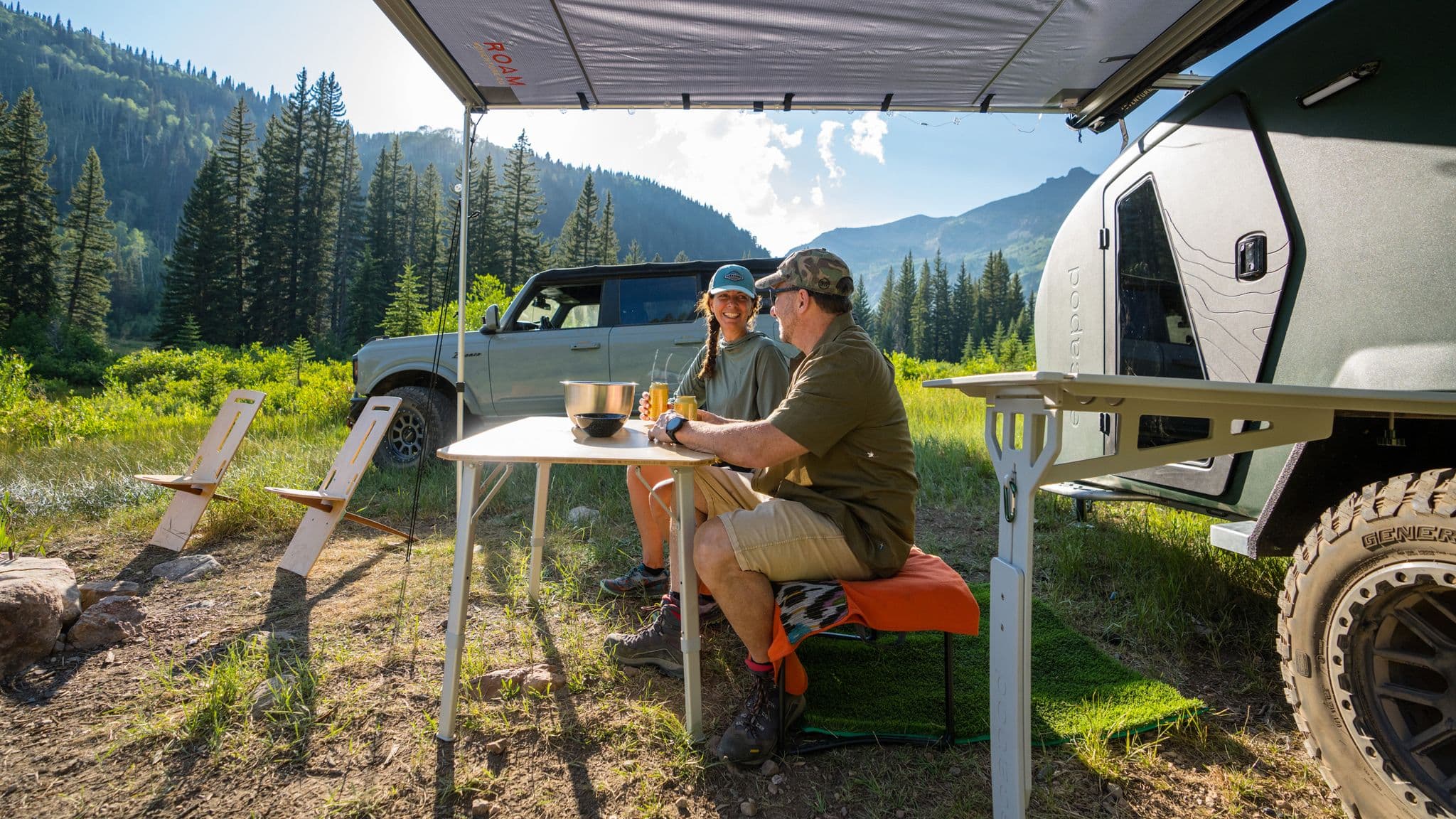 Couple enjoying a campsite with an Escapod trailer, foldable chairs, and a scenic mountain backdrop.