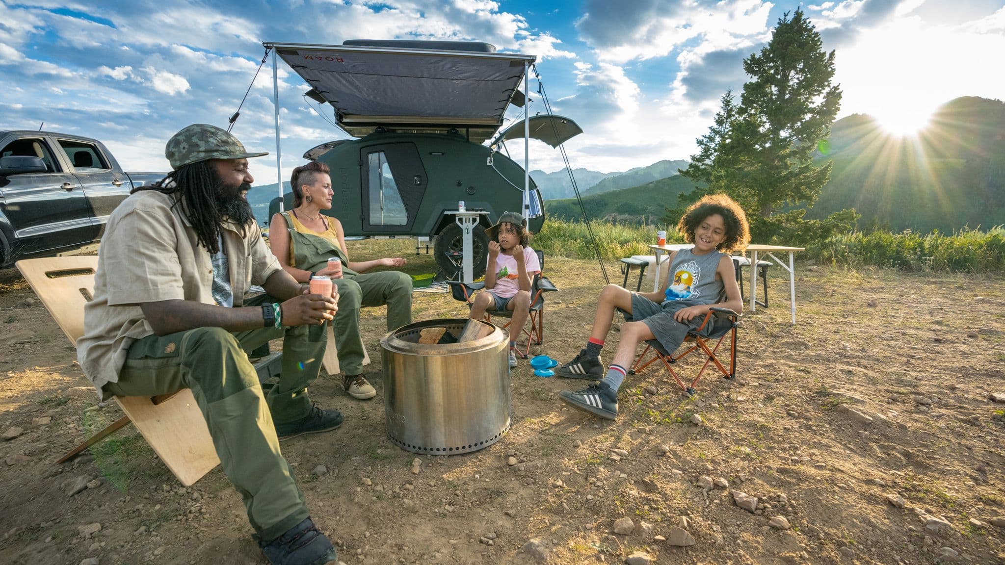 Family relaxing around a fire pit near an Escapod trailer, set up with an awning and a pickup truck, overlooking a mountain landscape.