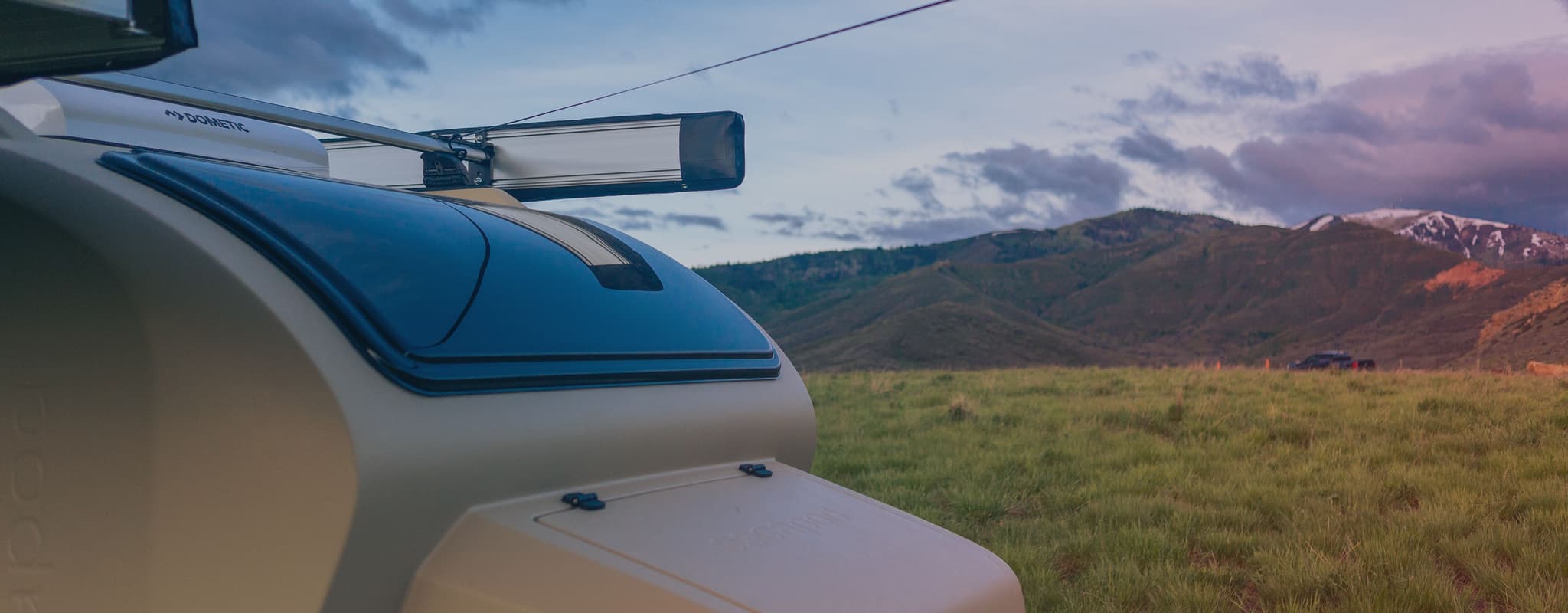 Close-up of an Escapod TOPO2 trailer’s skylight and roof rack against a backdrop of rolling green hills and a dramatic sky at sunset.