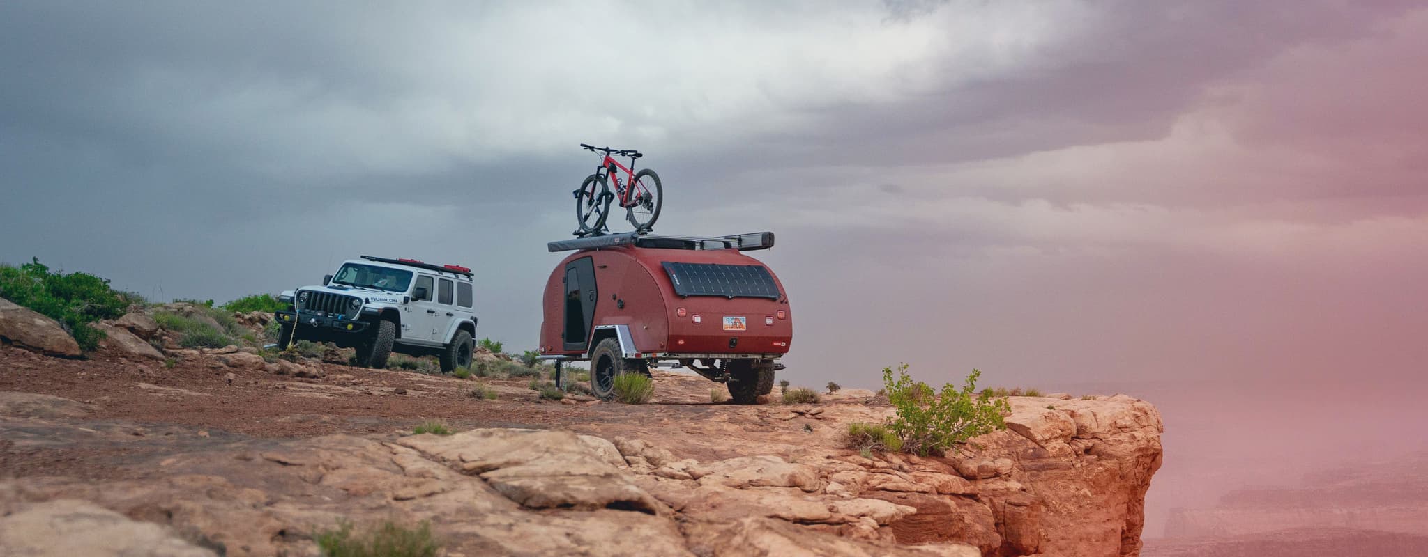 Rear view of an Escapod TOPO2 trailer with solar panels mounted on the back, parked on a desert cliff with a Jeep Wrangler and storm clouds overhead.