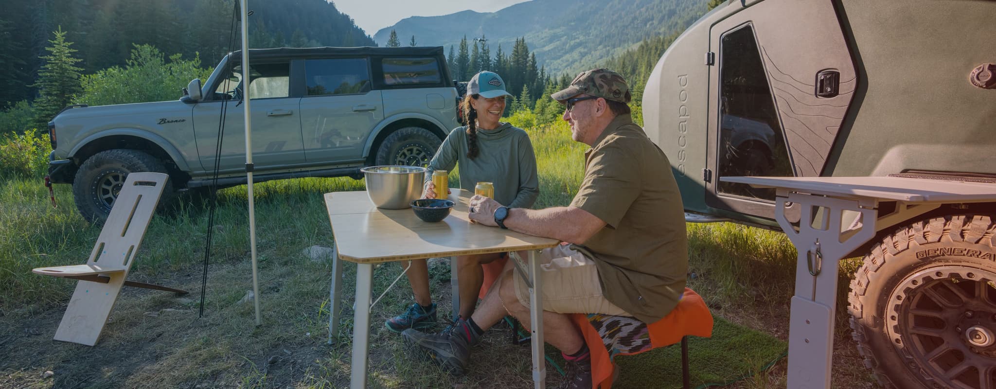 Man and woman sitting at a table under an Escapod trailer's awning, surrounded by nature.