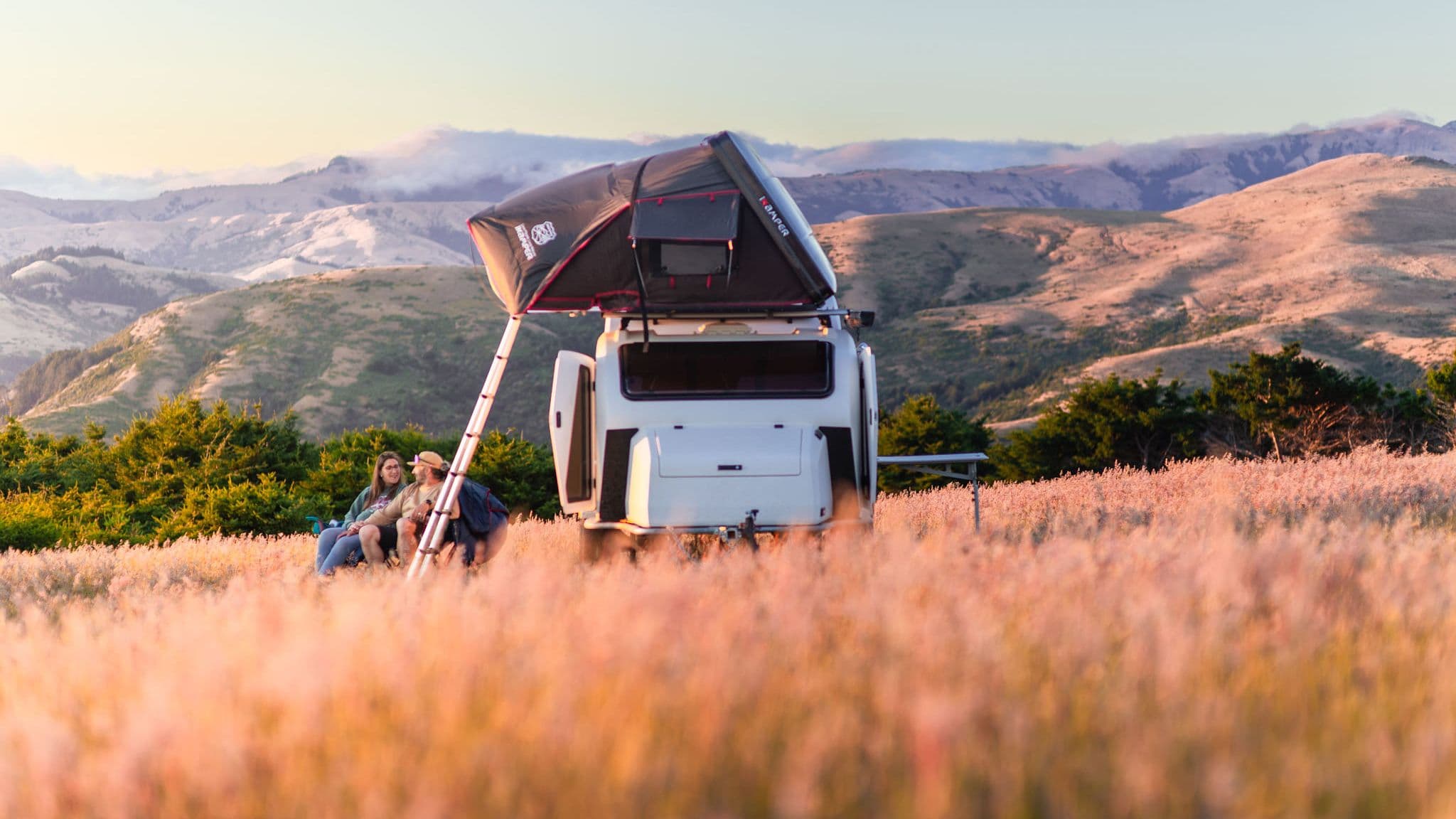 Two campers sitting in the grass near an Escapod TOPO2 with an iKamper tent, enjoying the sunset.
