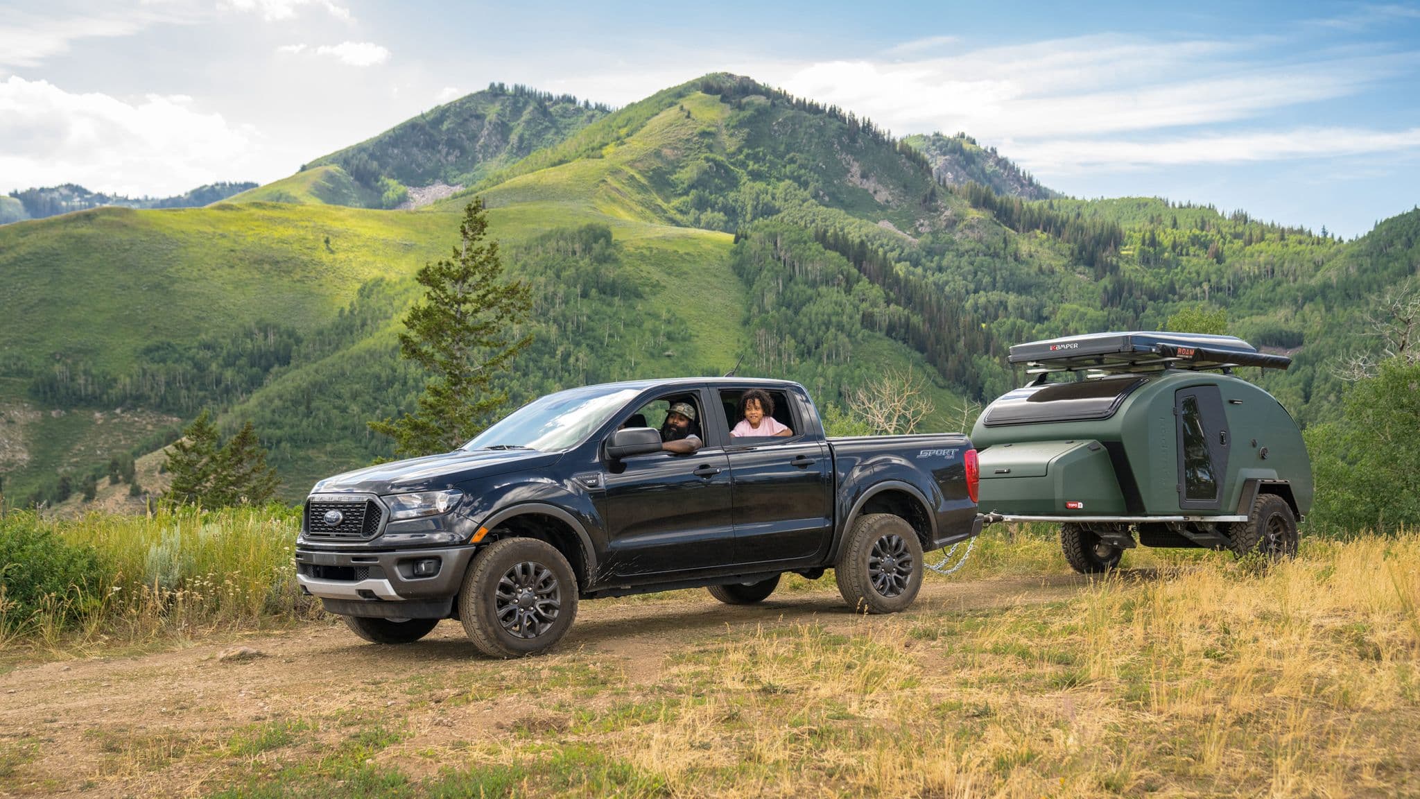 Ford Ranger towing an Escapod trailer through a mountain valley with green rolling hills, highlighting its compact, off-road camping capabilities.