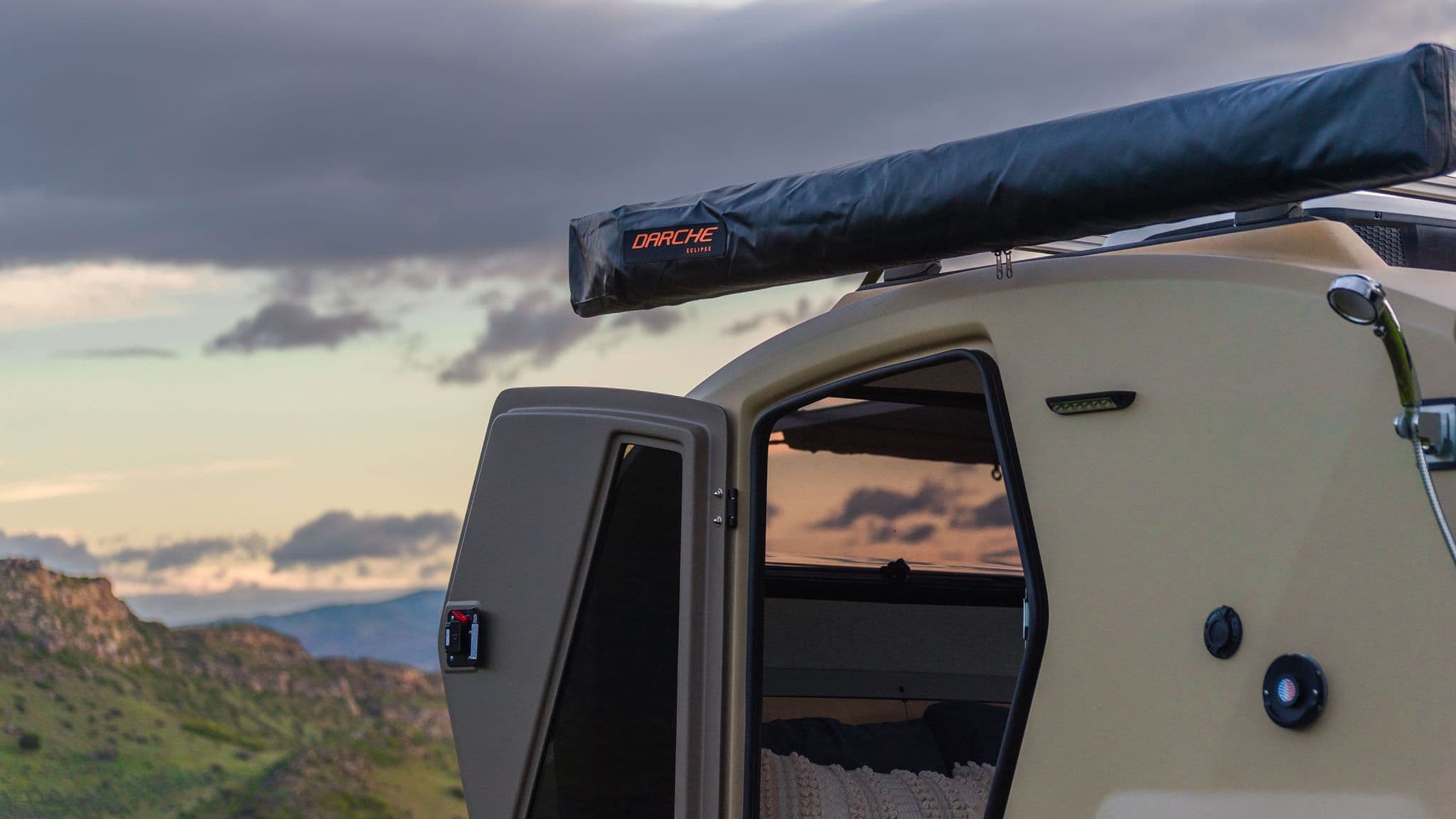Close-up of the Escapod TOPO2 trailer with door open at sunset, showing cozy interior bedding and mounted Darche awning for shade.