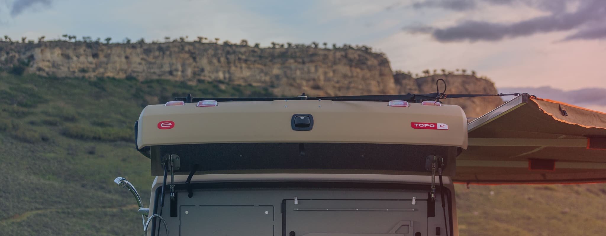 Rear view of an Escapod TOPO2 trailer with its galley open, set against a rugged landscape with cliffs and a cloudy sunset.