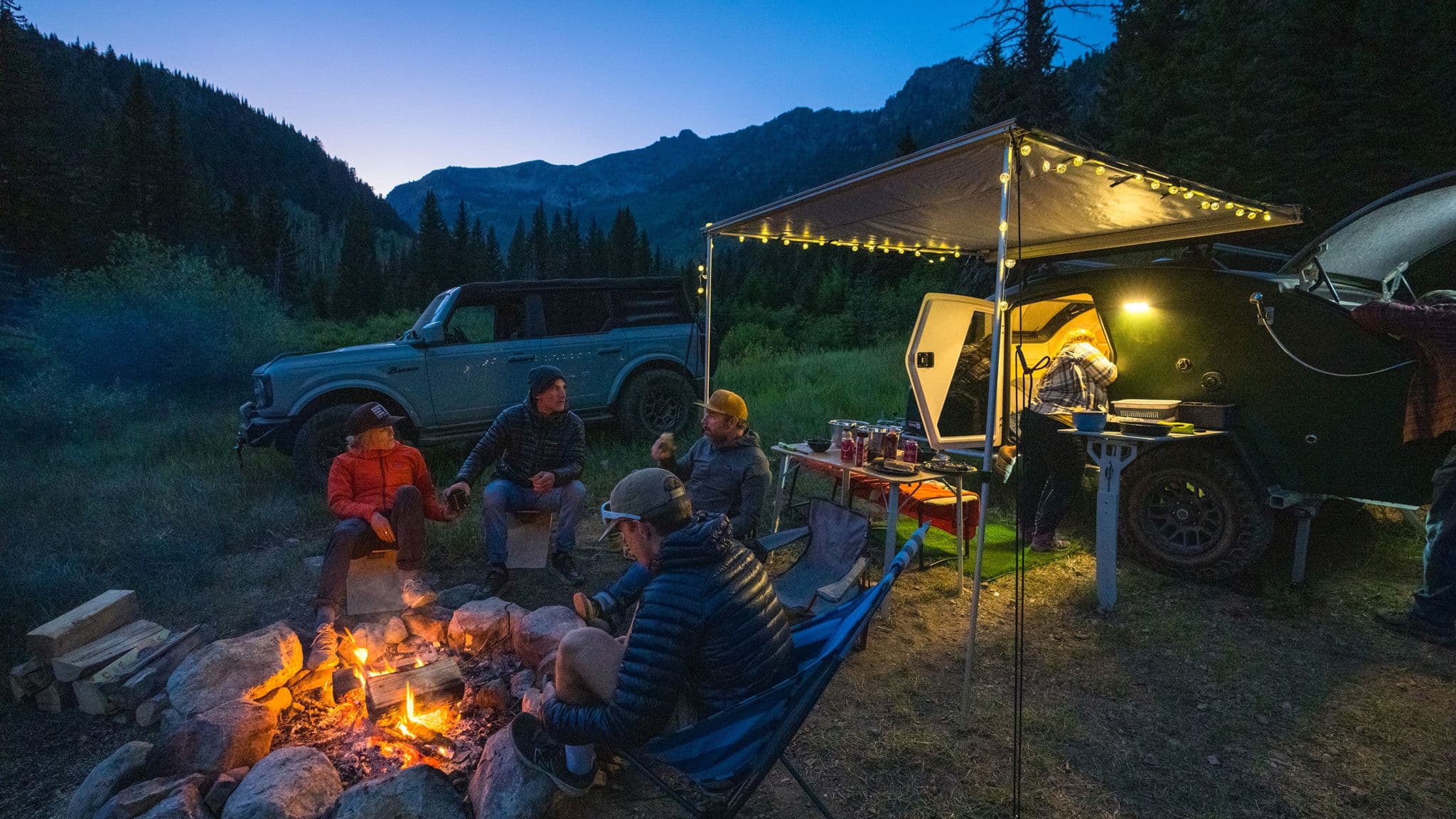 Group of campers relaxing by a fire at dusk with an Escapod trailer under string lights, cooking in the galley while surrounded by mountain views.