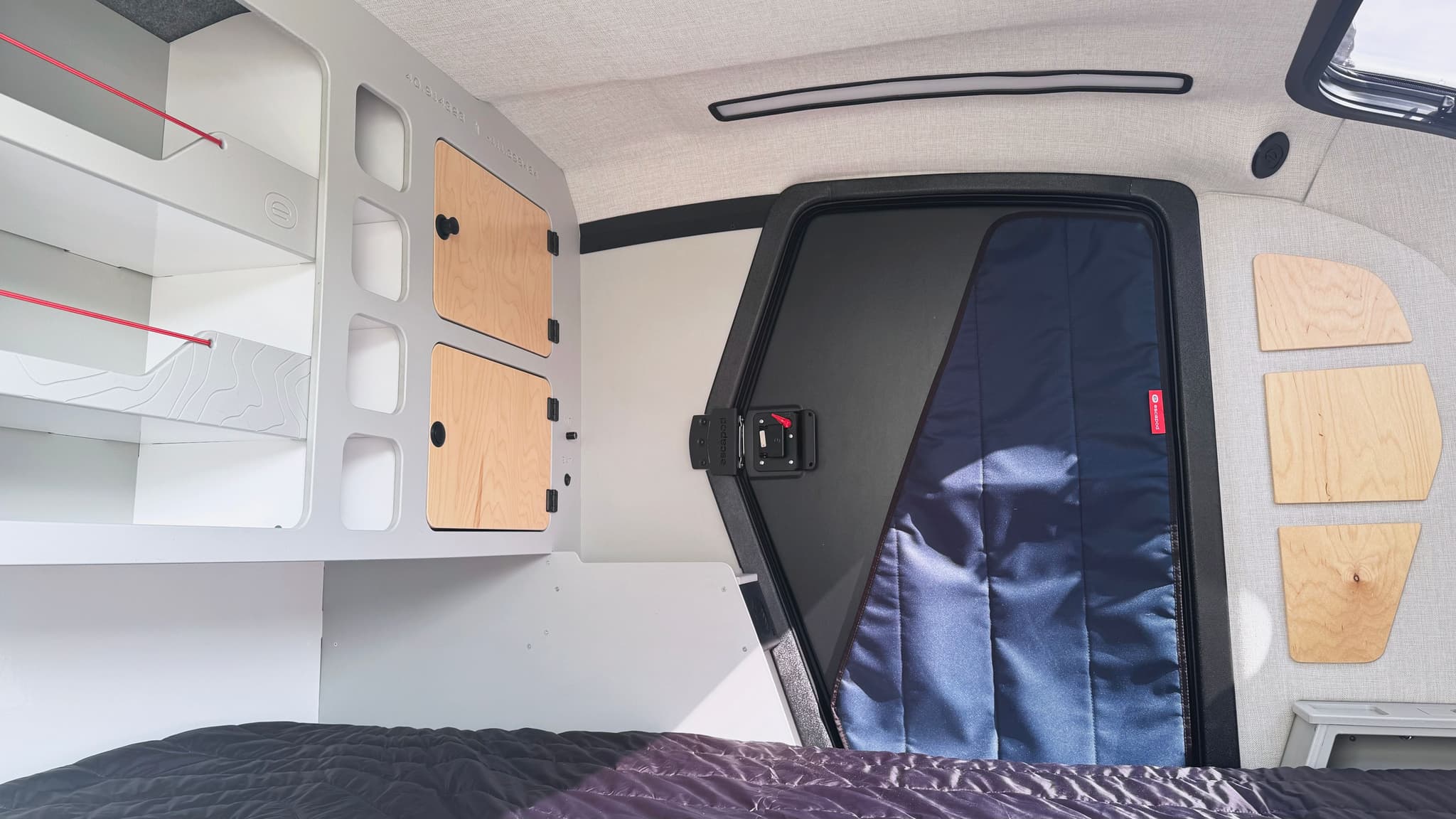 A wide angle view of the interior of an Escapod TOPO2 Nomad, showing cabinets, cubbies, open shelving, and a black entry door with a blue blackout window cover.