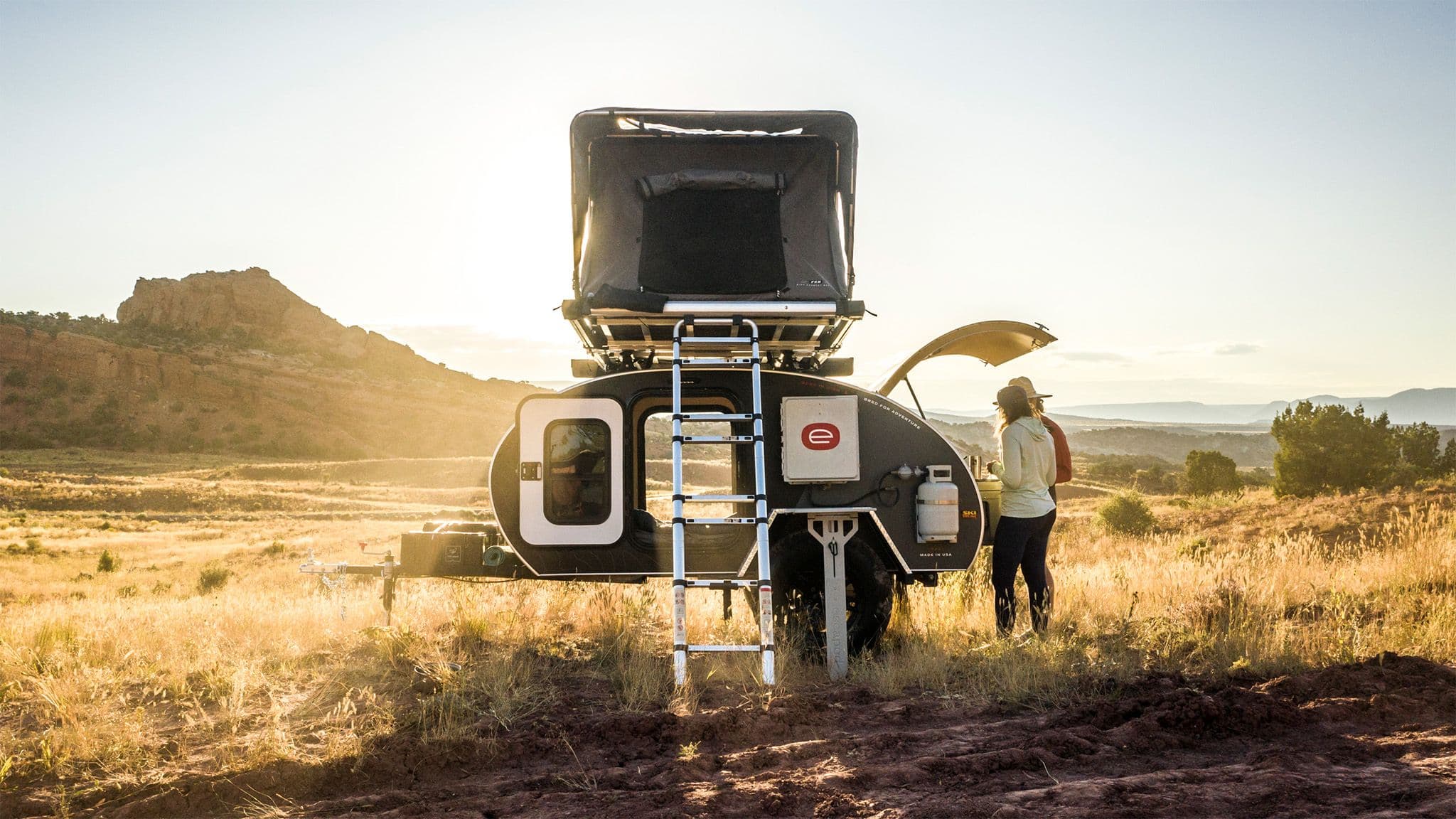 An Escapod TOPO2 trailer set up for camping in a golden desert landscape, with a rooftop tent and two people preparing a meal at the galley.