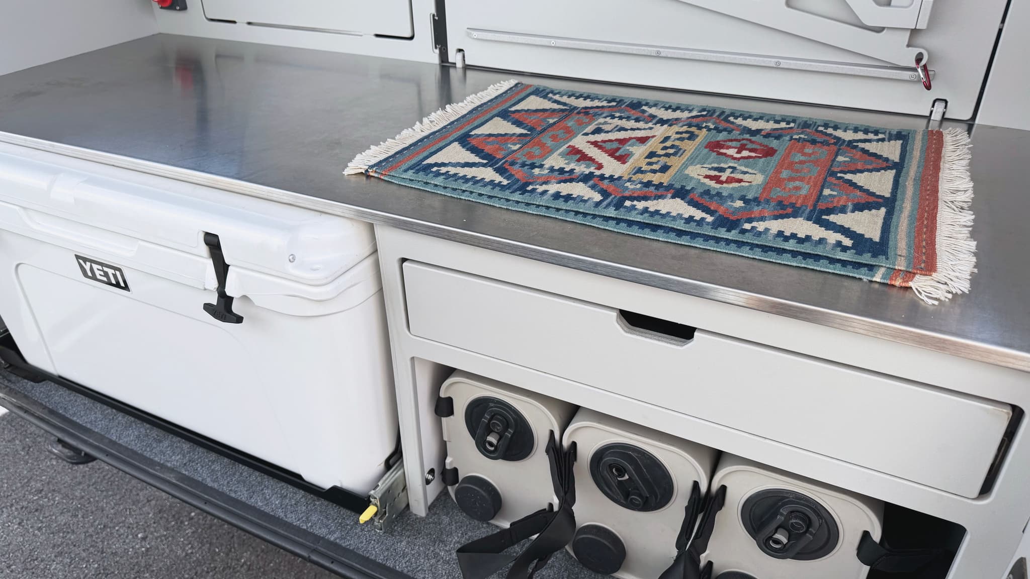 A wide angle view of the Escapod TOPO2 Nomad galley, showing the wide stainless steel countertop with a small patterned rug on top of the counter, and YETI cooler and Dometic water containers stored below.