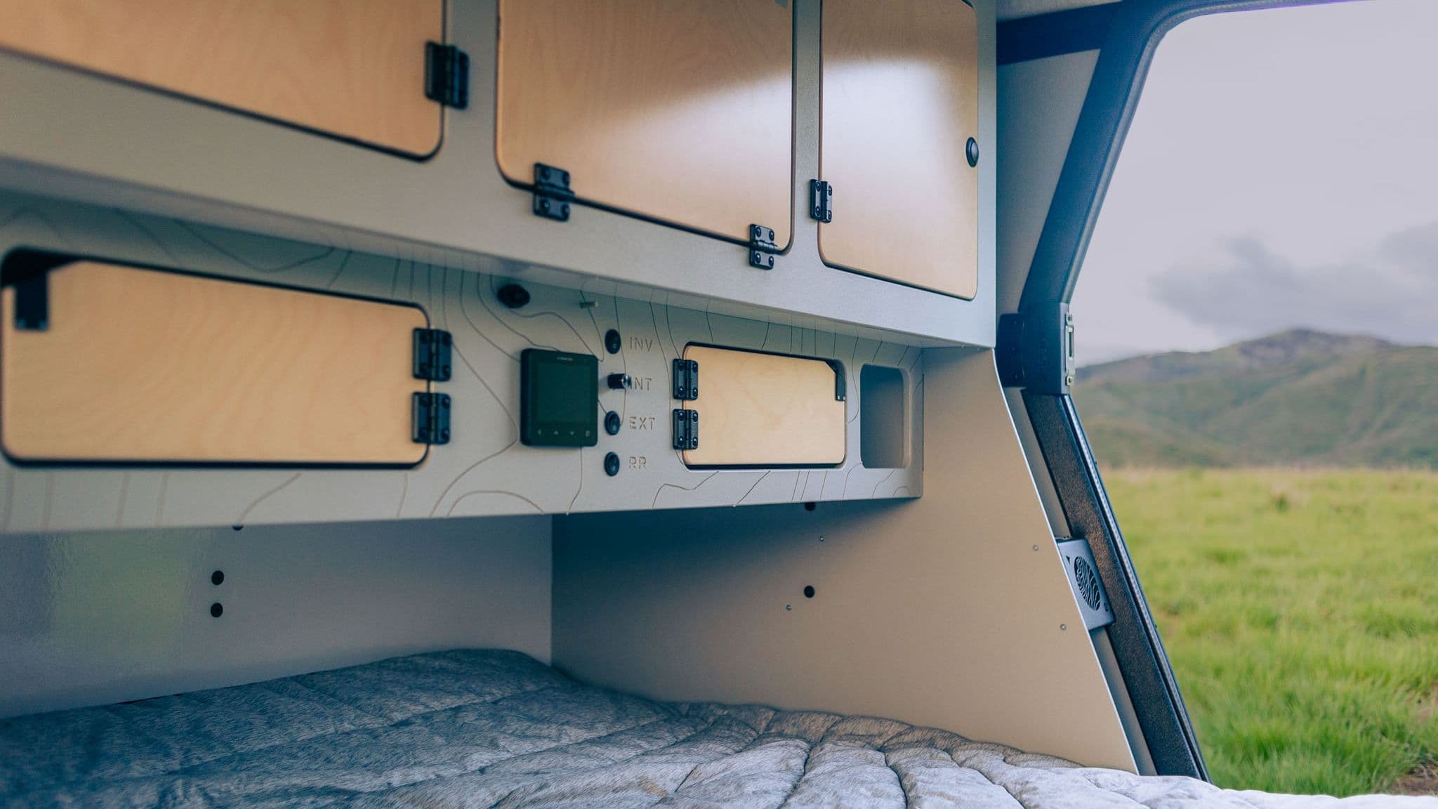 Interior view of the Escapod TOPO2, showcasing spacious birch cabinetry, soft bedding, and control panel, with an open door revealing lush green scenery.