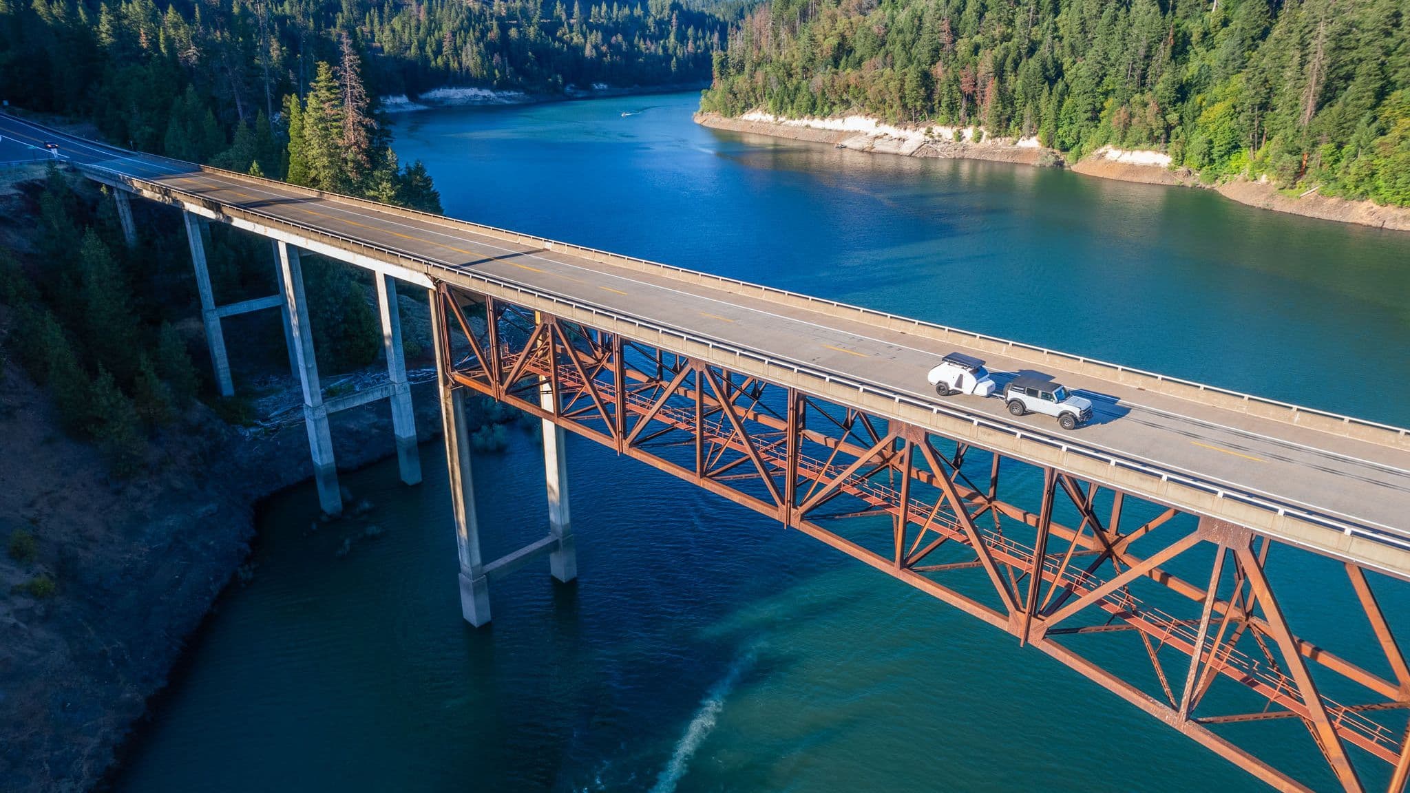 A Ford Bronco towing an Escapod trailer crosses a large steel bridge high above a deep blue river, surrounded by evergreen forests.