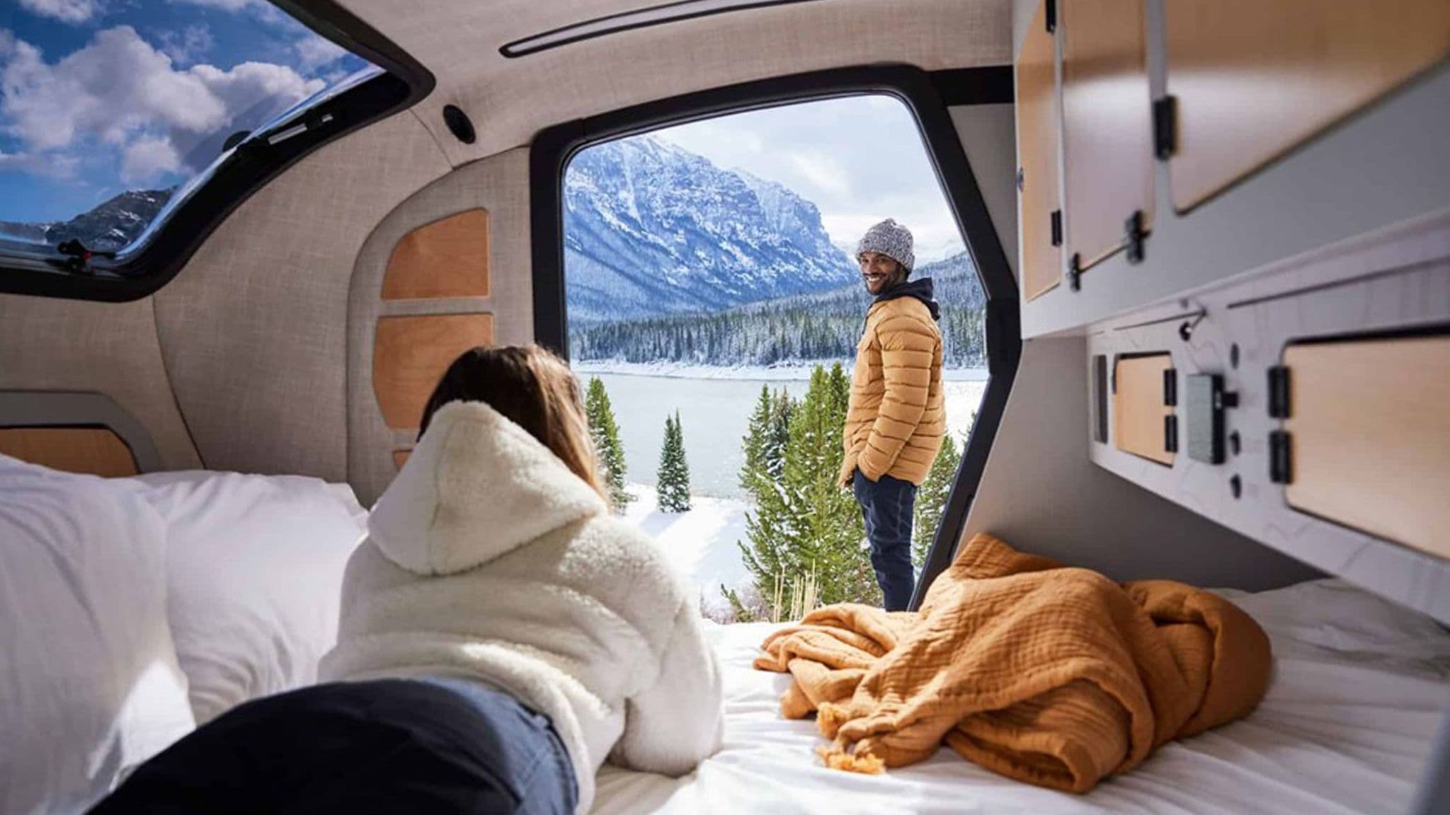 A woman relaxing inside an Escapod TOPO2 trailer, gazing at a man standing outside in a snowy mountain landscape through the large open door.